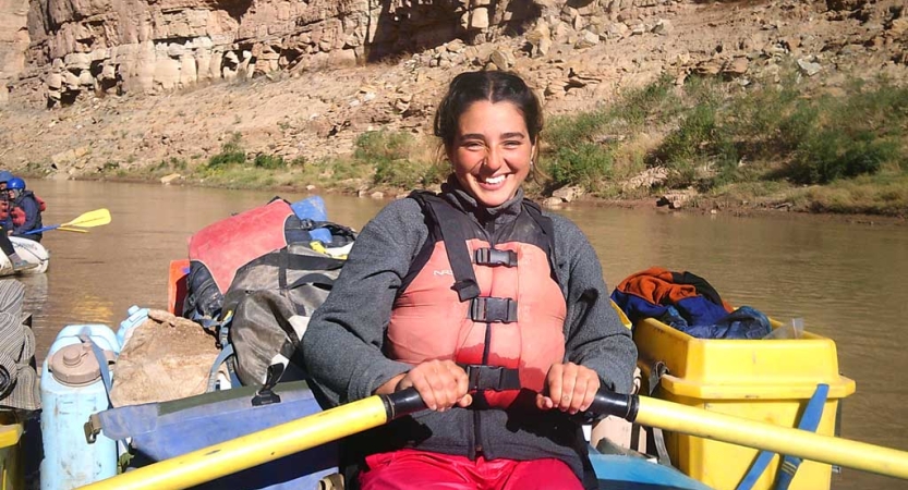 a person wearing a life jacket smiles as they hold the oars of a raft. Behind them is gear, the calm river and tall rock walls. 